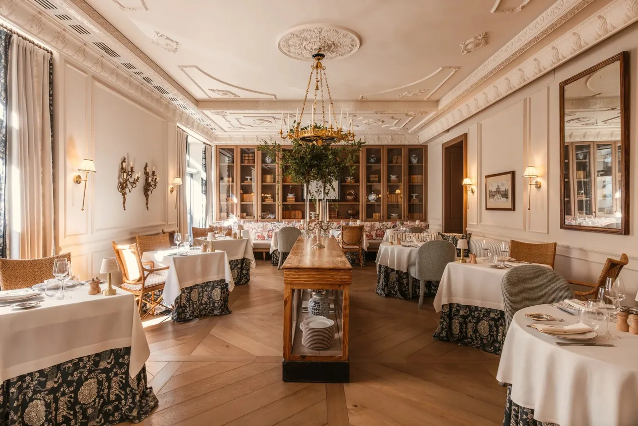 A large dining room with a high ceiling, white-clothed tables, a chandelier and bookshelves along the back wall