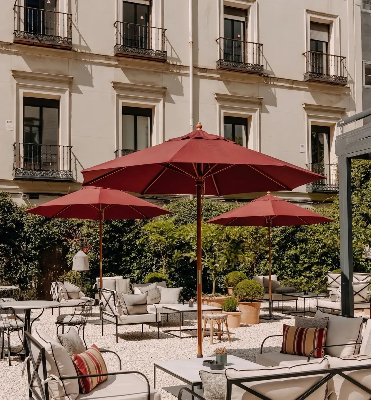 Red parasols over outdoor seating on a gravel courtyard terrace in front of a building with balconies