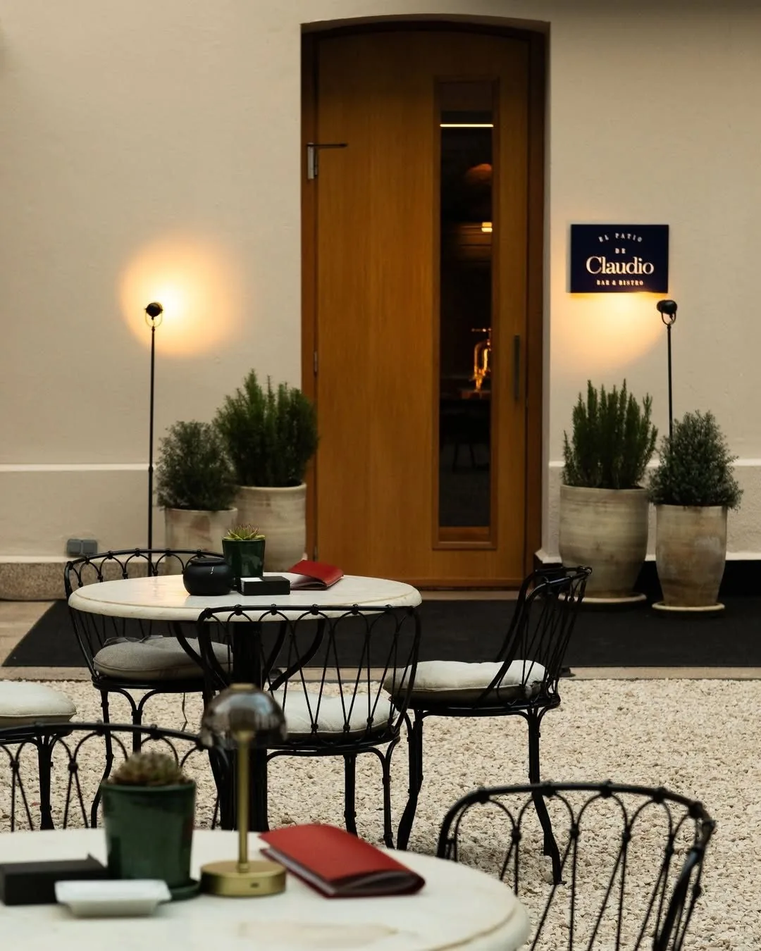 Round outdoor tables and black metal chairs by a wooden door on a gravel patio