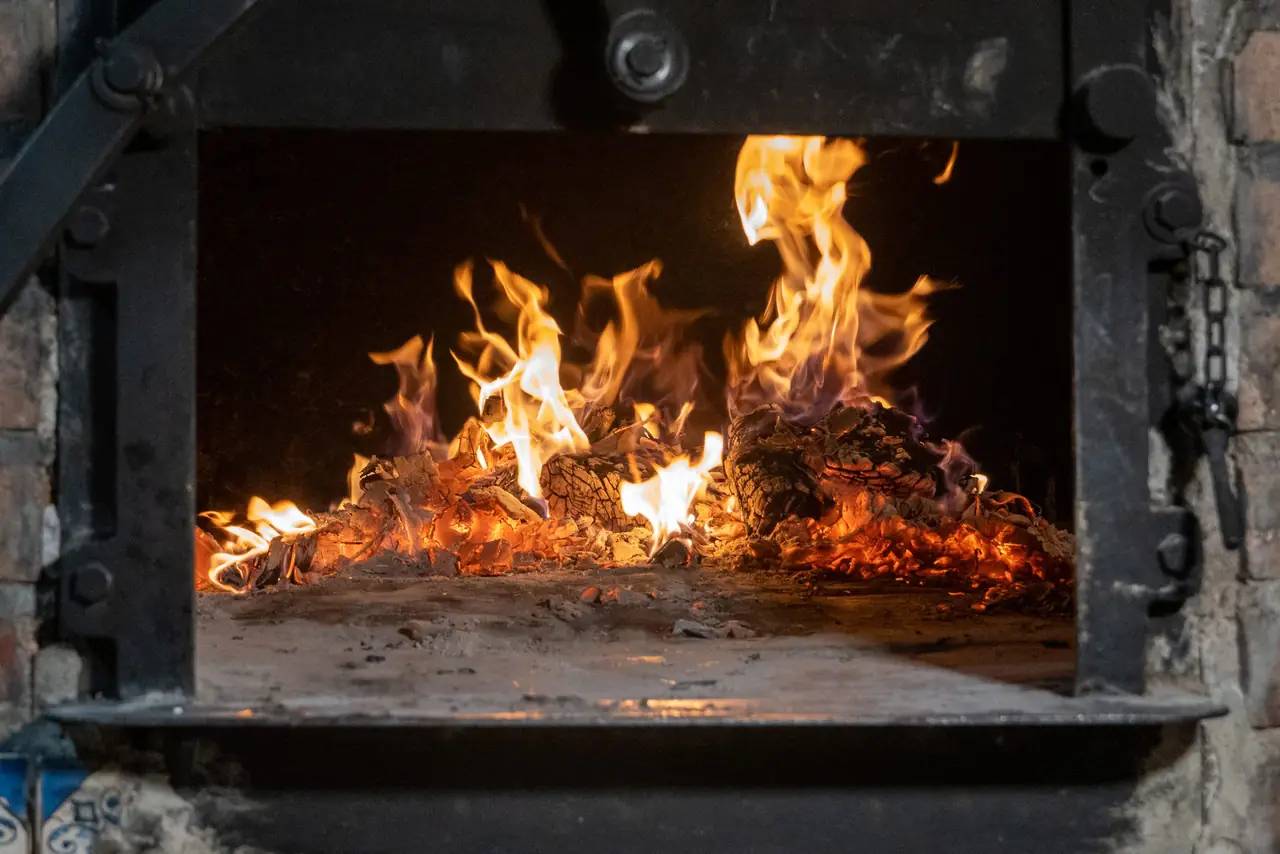 Wood fire burning inside the open mouth of the restaurant's iron-framed brick roasting oven, logs and embers glowing on the hearth floor with blue-and-white tiles at the edge