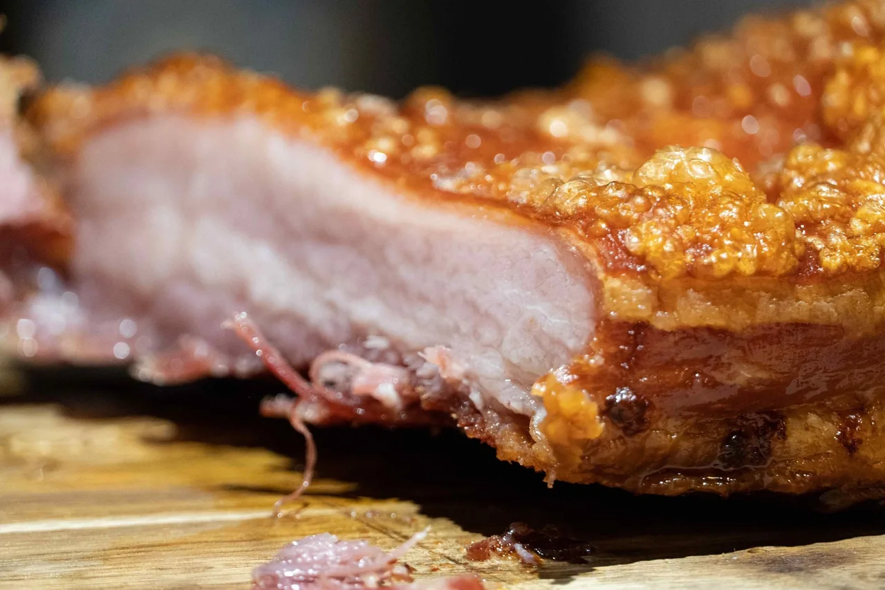 A close-up of a slice of roast pork with pink meat and blistered golden crackling on a wooden board