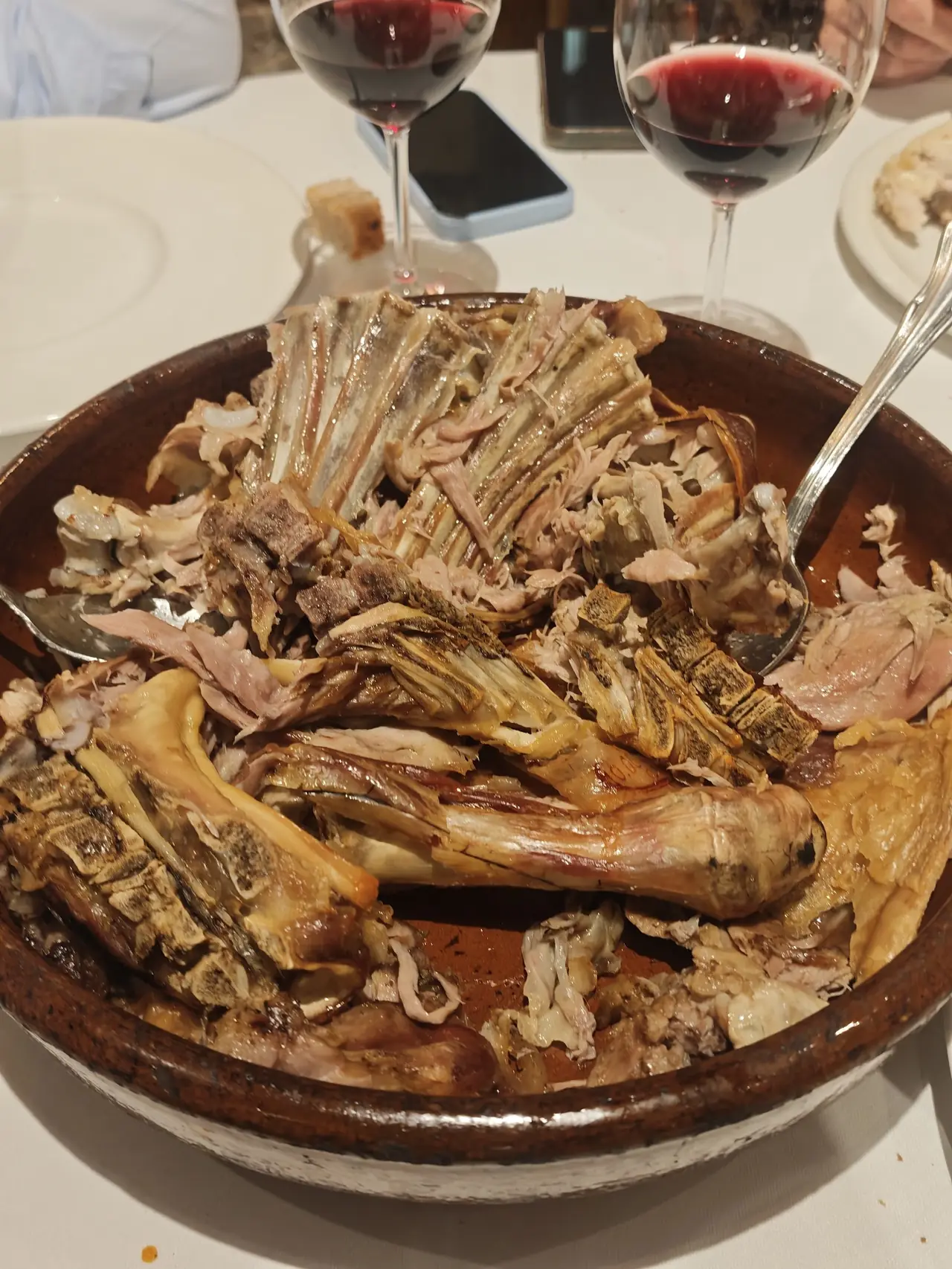 A large earthenware dish of roasted meat ribs and bones on the table with glasses of red wine
