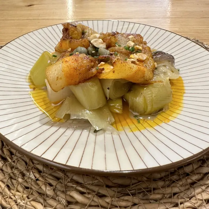 Crispy pan-seared fish over braised leeks with capers and chopped nuts on a striped plate at El Provisional