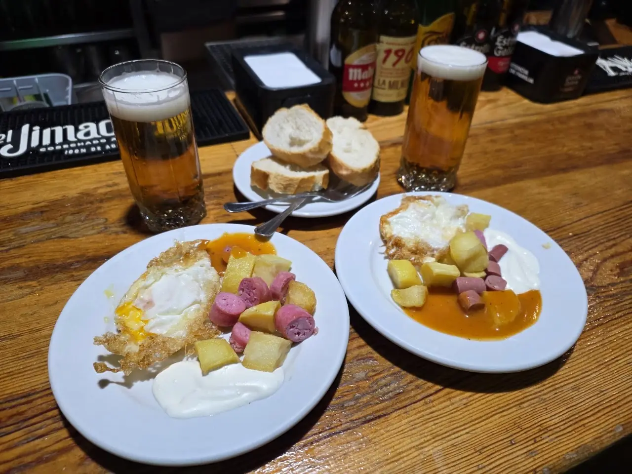 Two plates of fried eggs with potatoes and sliced sausage, with bread and two beers on the bar