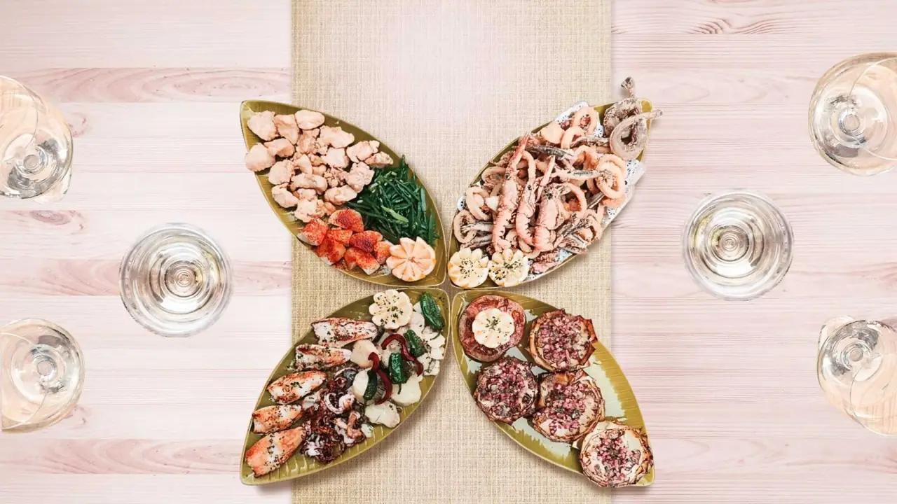 Four leaf-shaped platters arranged in a flower of fried and grilled seafood - langoustines, squid, prawns - framed by white wine glasses