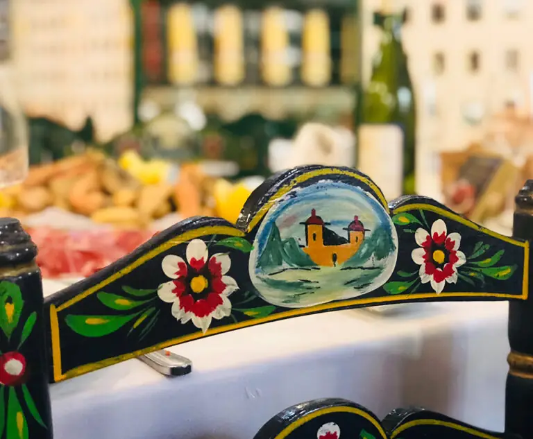 Close-up of a hand-painted black rush chair back with floral motifs and a village scene, a laid table of food blurred behind