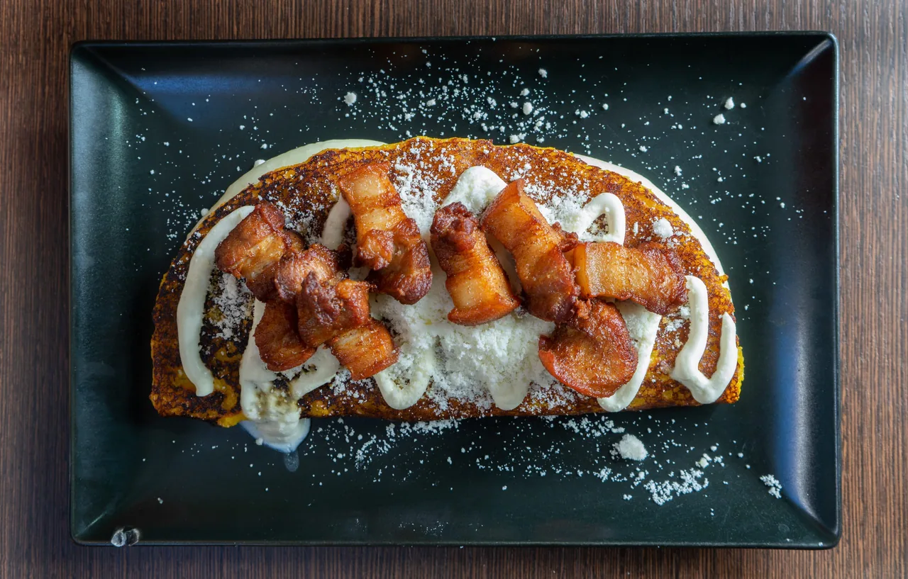A cachapa folded over melting white cheese and draped with strips of fried pork belly, dusted with crumbled queso, on a black rectangular plate on a dark wooden table