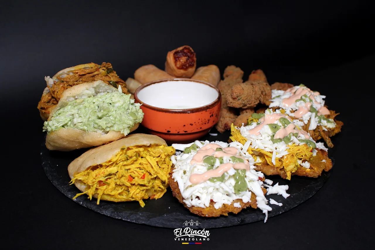 A slate board of Venezuelan snacks: arepas split and filled with shredded chicken and avocado salad, tequenos and fried pastries behind, and patacones topped with shredded chicken, white cheese and pink and green sauces, around a terracotta bowl of dip