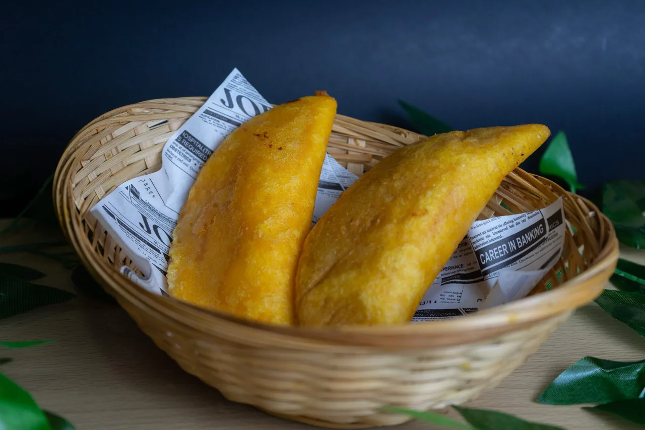 Two deep-fried golden empanadas resting in a woven basket lined with newsprint-patterned paper, against a dark background with green leaves