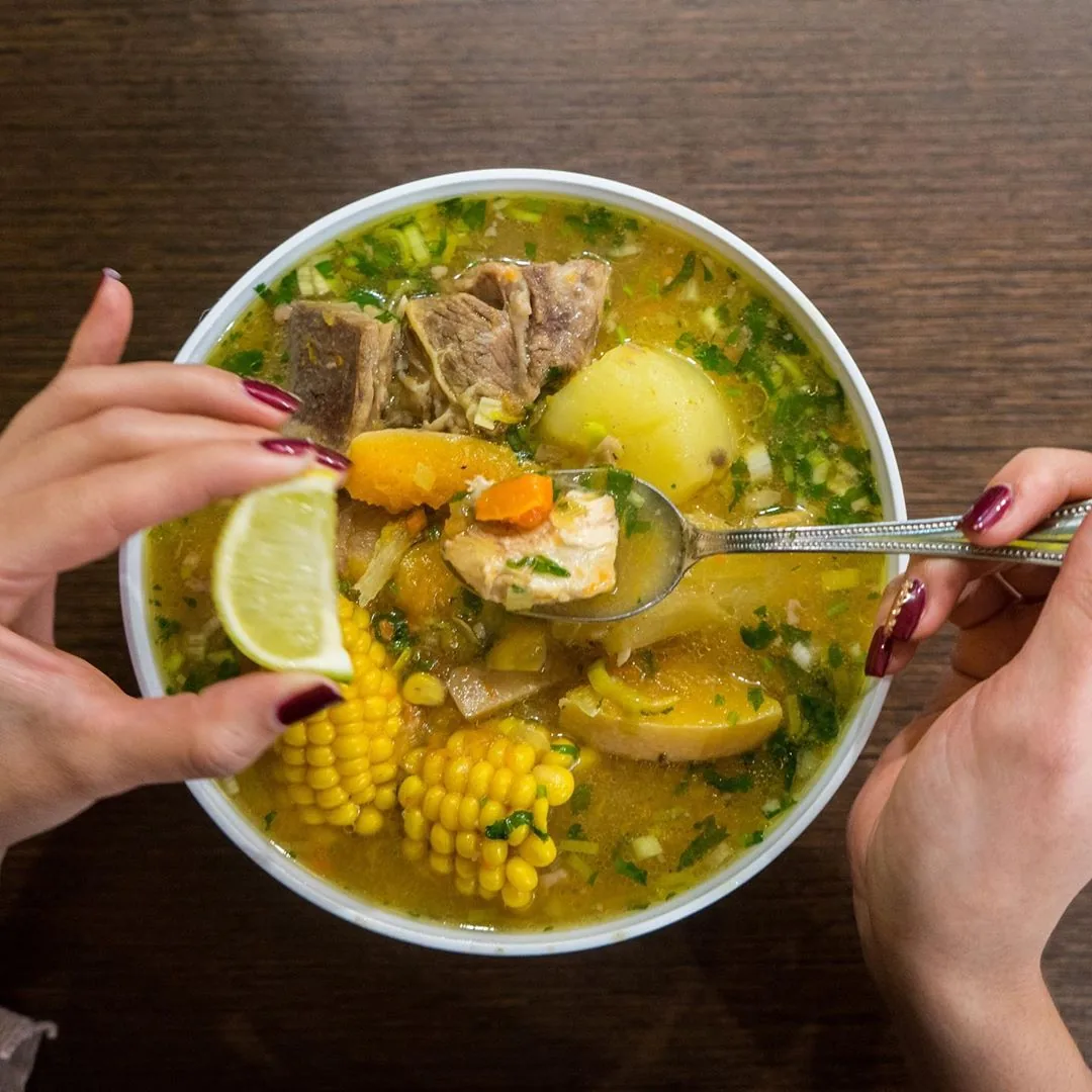 Two hands over a white bowl of sancocho, clear broth with a rib of meat, potato, pumpkin, corn on the cob and chopped herbs, one lifting a spoonful, the other holding a lime wedge