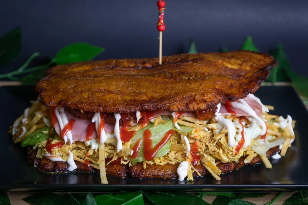 A patacon sandwich built between two large fried plantain slices, filled with shredded chicken, ham, lettuce and shoestring potatoes and laced with ketchup and white sauce, pinned with a beaded skewer on a black plate
