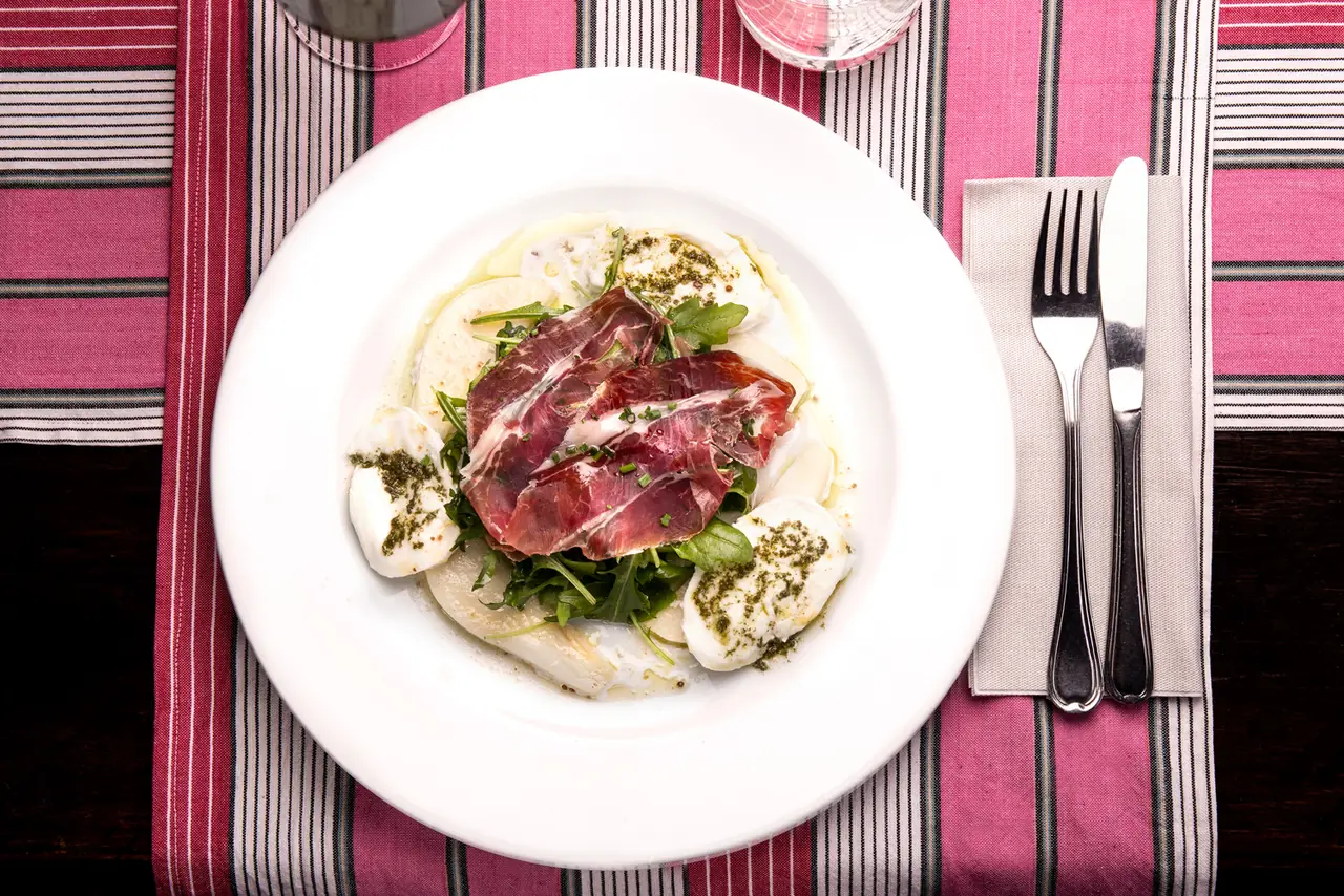 Sliced Iberian ham draped over burrata-style mozzarella and arugula, drizzled with herb oil, on a white plate at El Salón