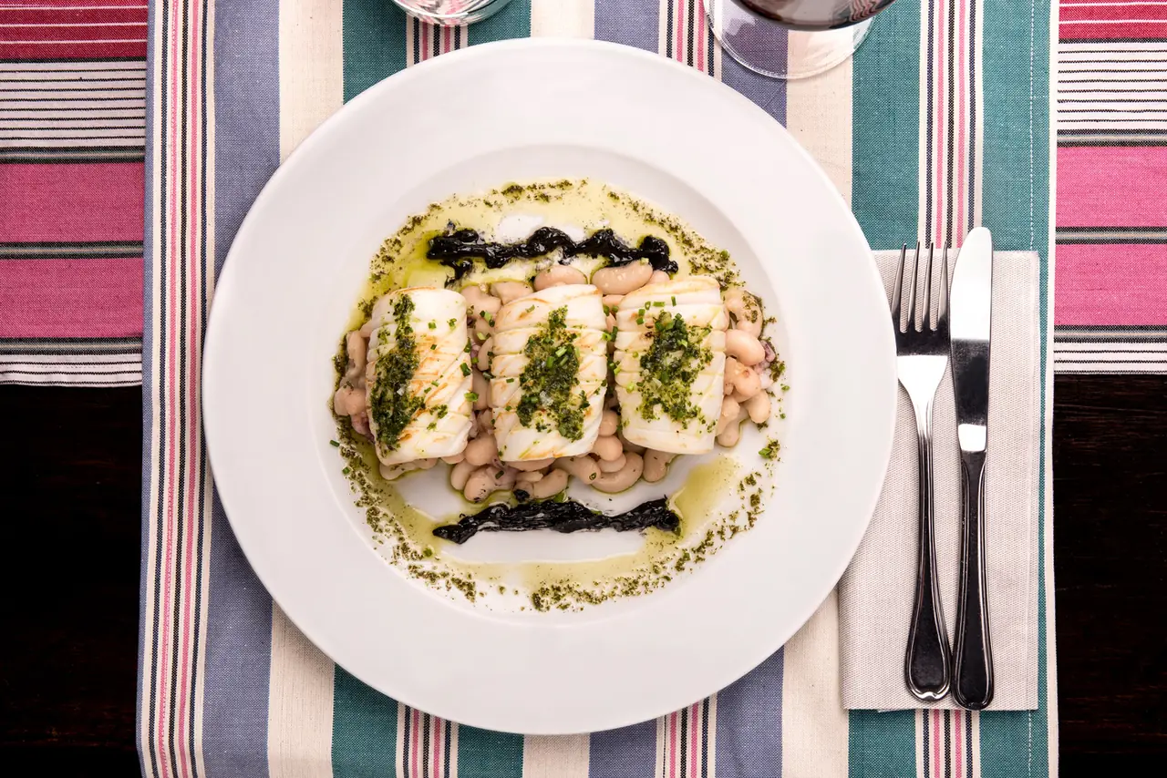 Seared white fish fillets over white beans with parsley oil and squid-ink accents on a white plate at El Salón