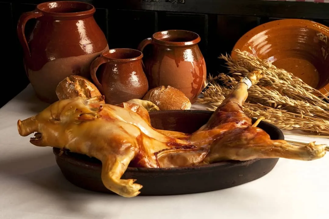 A whole roast suckling pig in a dark dish beside earthenware jugs and bread rolls