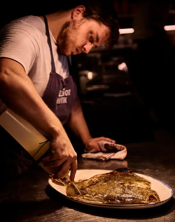 In a dim kitchen a cook in a dark apron embroidered with the restaurant's name leans over a steel counter and spoons pale sauce from a bottle over a whole grilled flatfish resting on a blue-rimmed enamel platter.