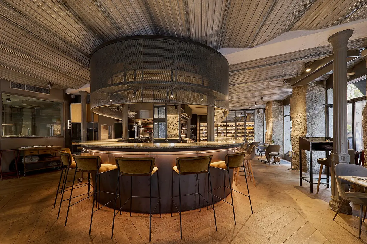The circular central bar counter with olive-green upholstered stools all round it under a wide mesh drum canopy, herringbone parquet floor and stone columns.