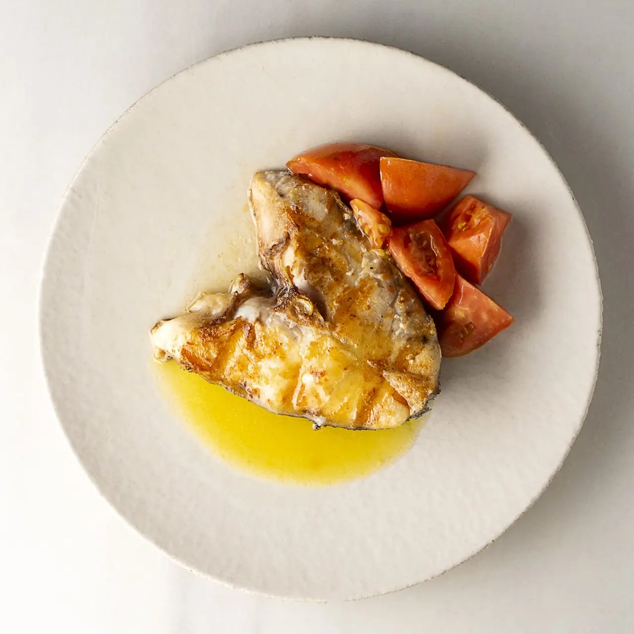 A grilled fish fillet served with fresh tomato wedges in olive oil on a white plate.