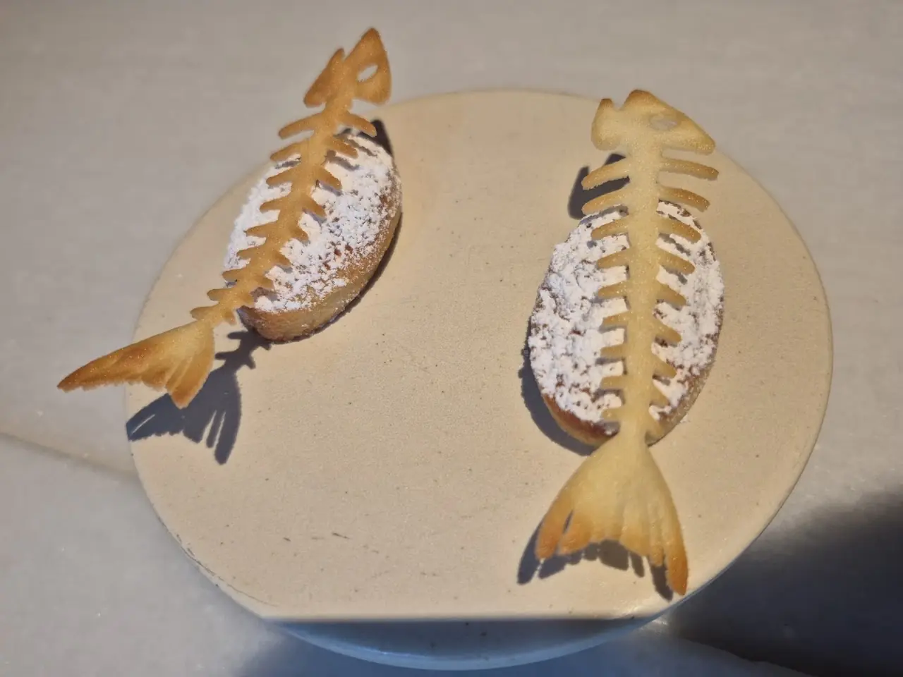 Two fish-bone-shaped pastries dusted with powdered sugar on a round plate.