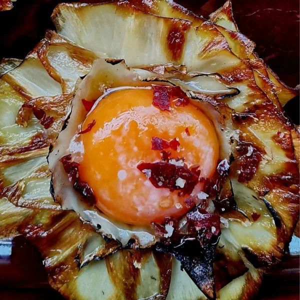 A glossy orange egg yolk sitting at the centre of a rosette of charred grilled mushroom slices, scattered with red chilli flakes and flakes of sea salt