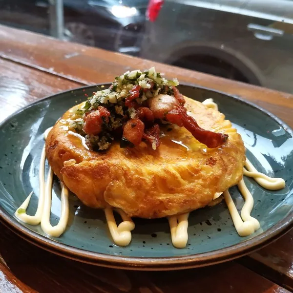 A thick golden tortilla on a speckled teal plate, topped with reddish octopus pieces and a green herb-and-crumb mix, ringed with piped dots of alioli, on a wooden table by a window