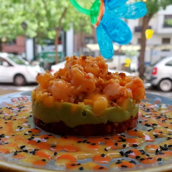 A layered timbale of prawns over avocado and diced tomato on a plate pooled with orange sauce and black sesame seeds, photographed against a sunlit street of parked cars and trees