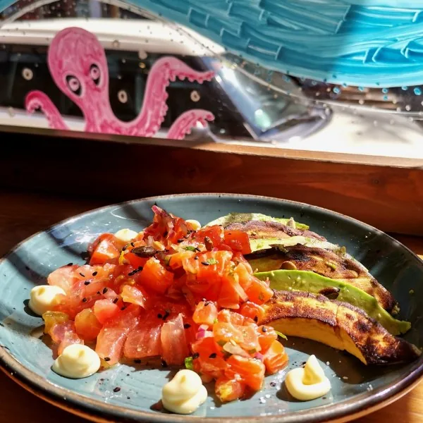 Diced raw fish and tomato salsa with a grilled half avocado, dots of mayonnaise and black sesame on a speckled teal plate, with a pink octopus decal on the window behind