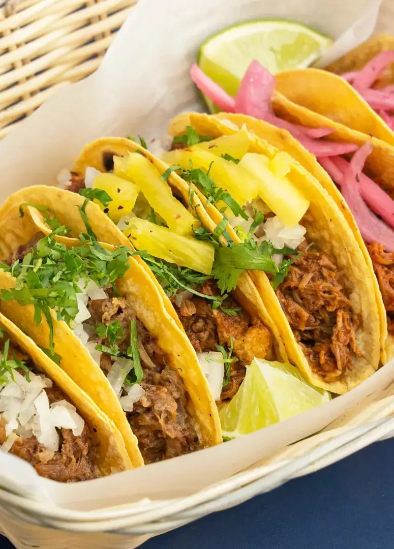 A basket of four folded corn tortilla tacos topped with shredded meat, chopped onion, cilantro, pineapple strips, and pickled red onion, with lime wedges, at El Tianguis