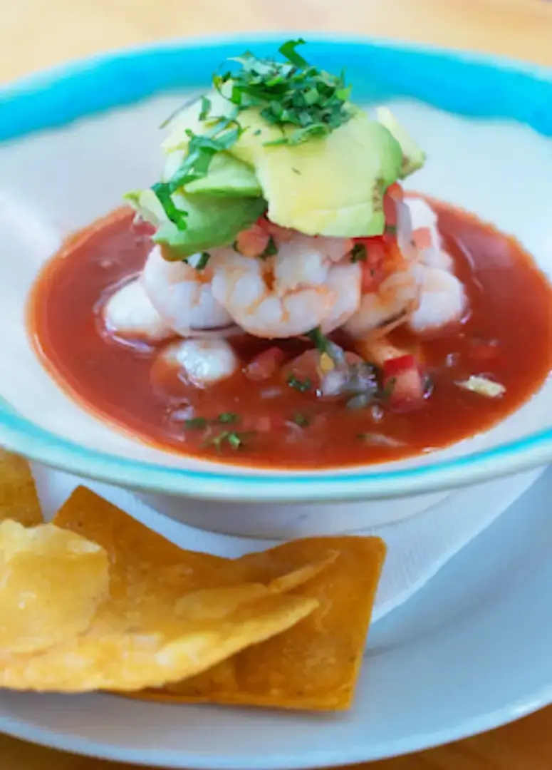 A shrimp ceviche in red tomato-based broth topped with sliced avocado and herbs, served in a blue-rimmed bowl with tortilla chips, at El Tianguis