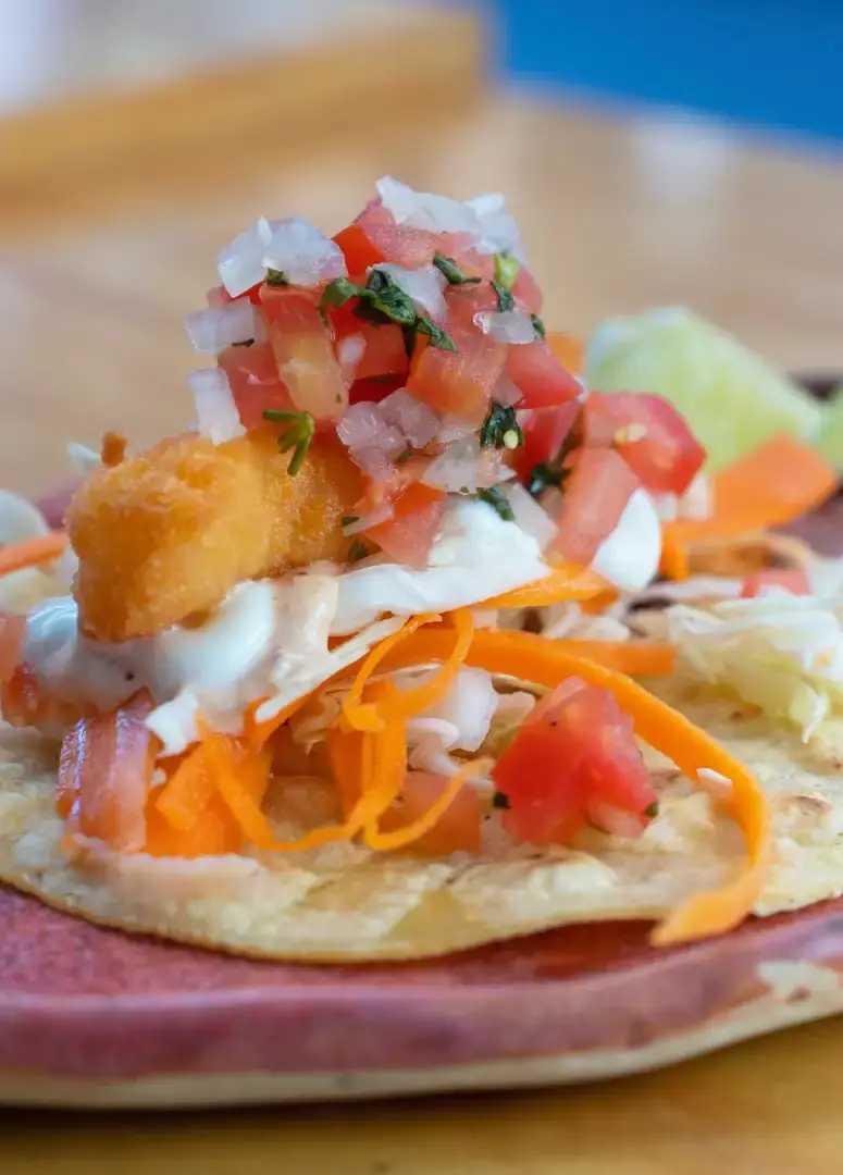 A tostada topped with fried fish, pico de gallo, shredded carrot, and cabbage on a corn tortilla base, at El Tianguis