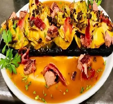 Plated dish of sliced meat and jamon over artichoke layers with brown sauce and parsley