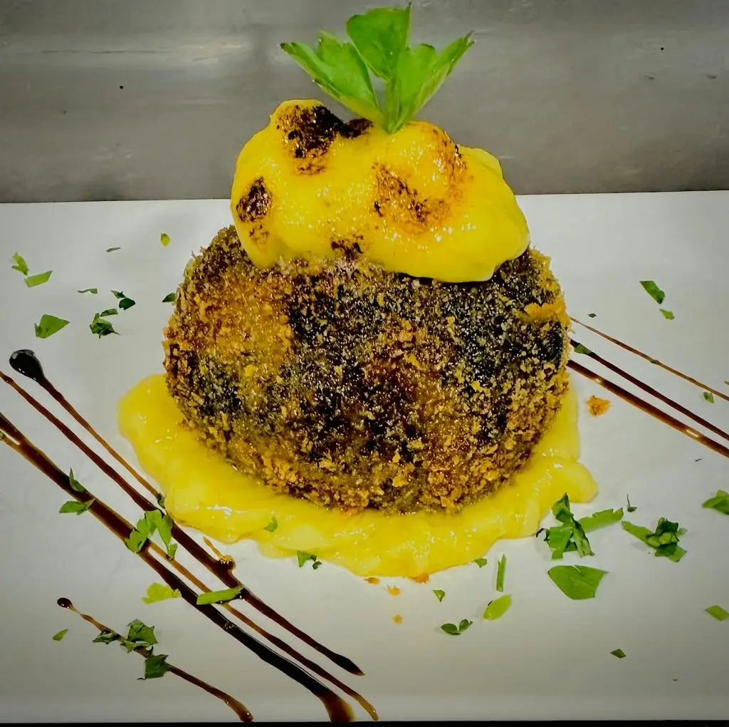 Breaded fried croqueta-style dome topped with torched yellow sauce and parsley, with balsamic drizzle on a white plate