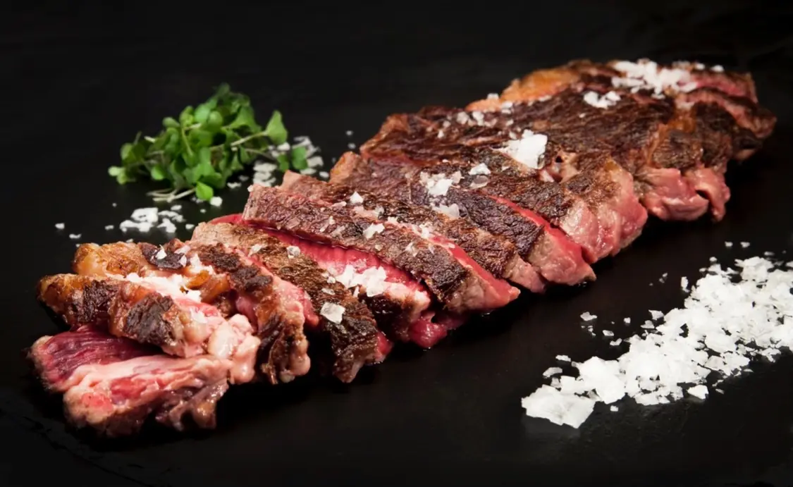 Sliced medium-rare beef entrecôte with flaky salt and microgreens on black slate