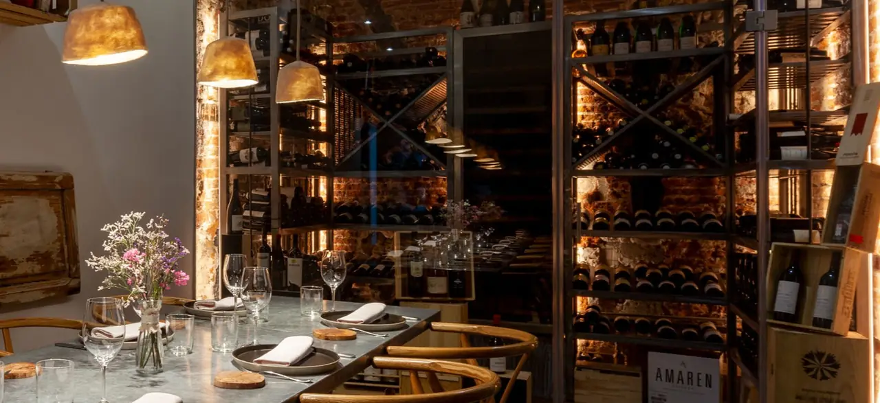 Table laid with glasses, napkins and small flowers beside a glass-fronted wine store, metal racks of bottles against exposed brick lit warmly, with gold pendant lamps overhead