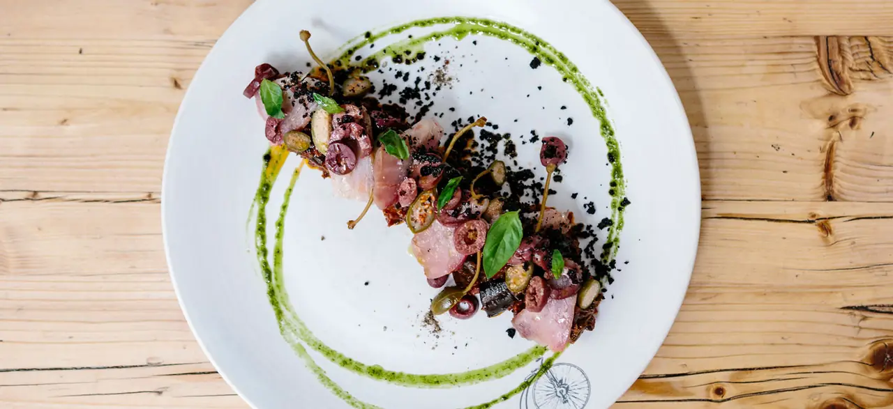 Overhead shot of thin slices of raw pink fish with olives, capers, sliced pickles, black crumb and basil leaves, ringed by a green herb-oil swirl on a white plate marked with a small bicycle-wheel drawing, on a bare pine table