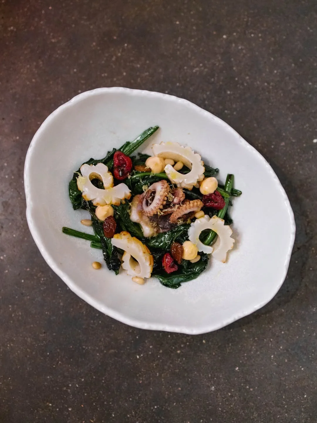 A white bowl of a salad of small seafood pieces with dark leaves and red pieces on a dark surface