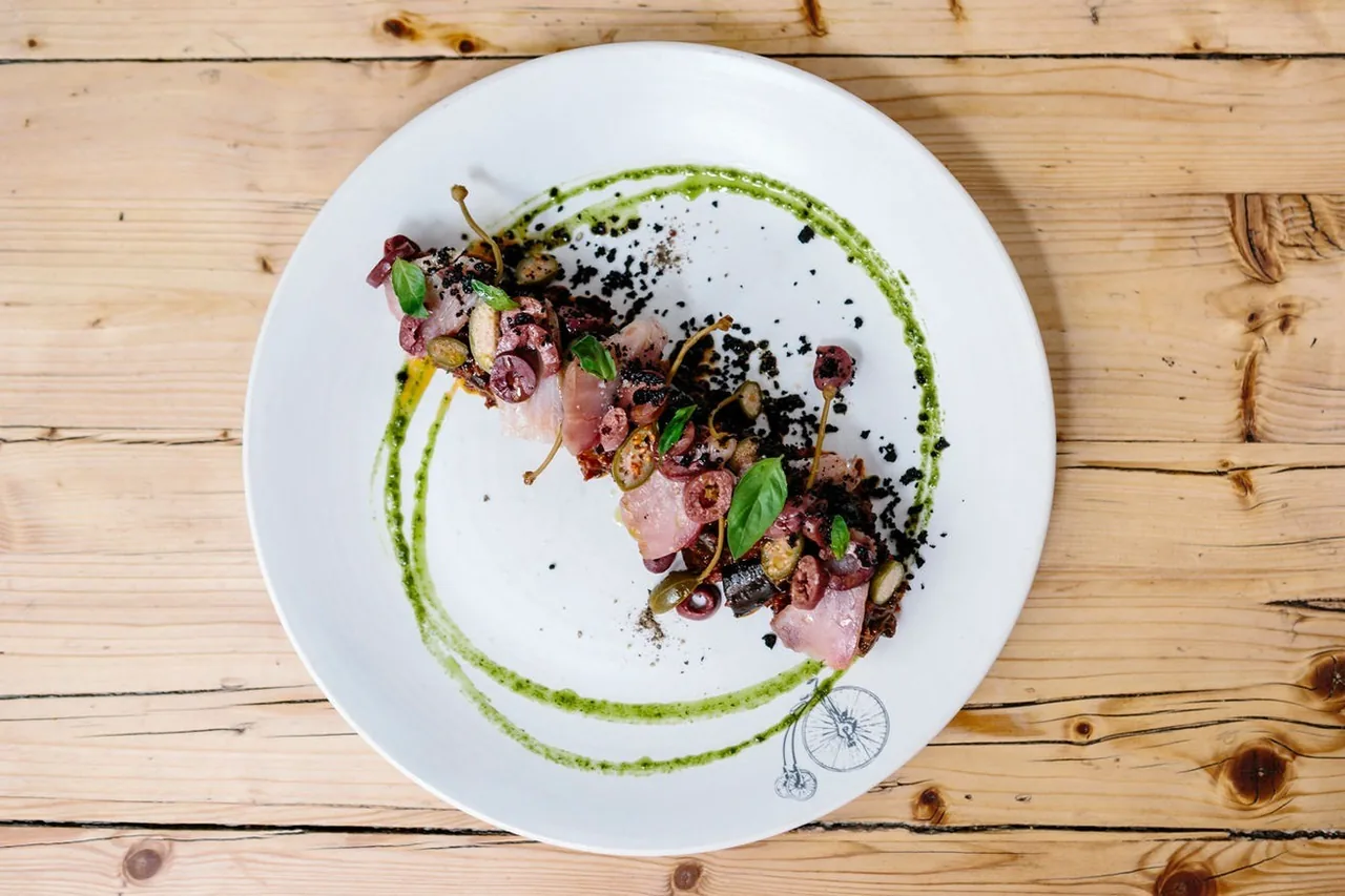 A white plate of sliced meat with dark crumbs and a green sauce ring on a wooden table