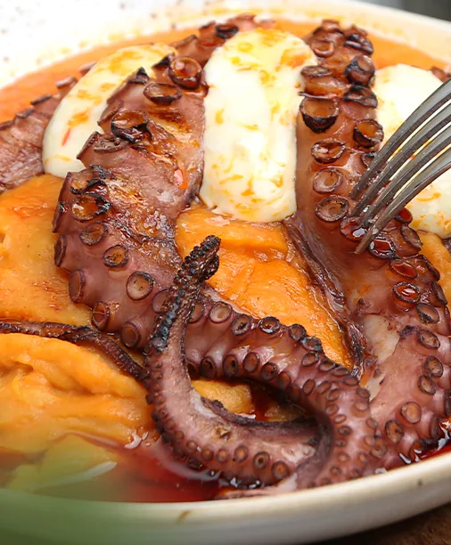 Octopus tentacles glossed with paprika oil over potato puree in a shallow ceramic dish, a fork resting at the edge