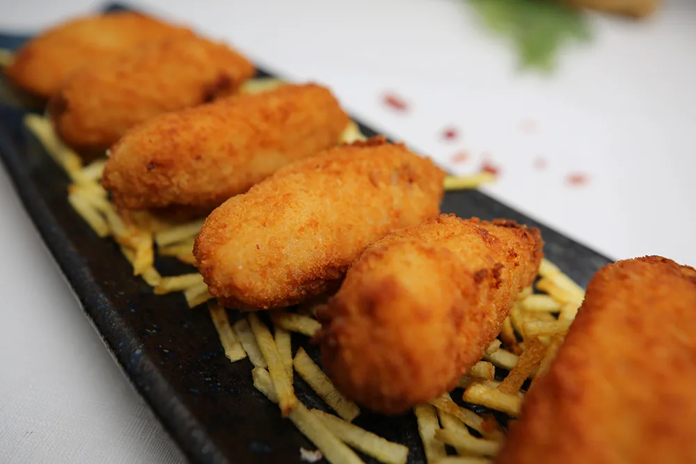 Five golden croquetas lined up on a dark slate board over a bed of matchstick fried potatoes