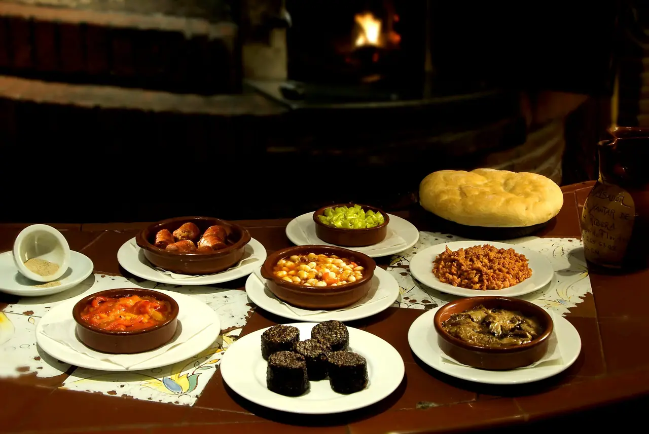 Spread of Castilian asador starters on a wood table by the fire — chorizo, white beans, roasted red peppers, morcilla rounds, pisto, braised mushrooms, and bread, El Yantar de la Ribera