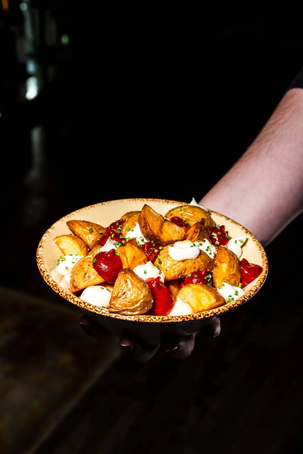 Bowl of crispy roasted potato wedges topped with dollops of aioli, red sauce, and chopped chives, held out on a black tabletop, at Eleven BCN