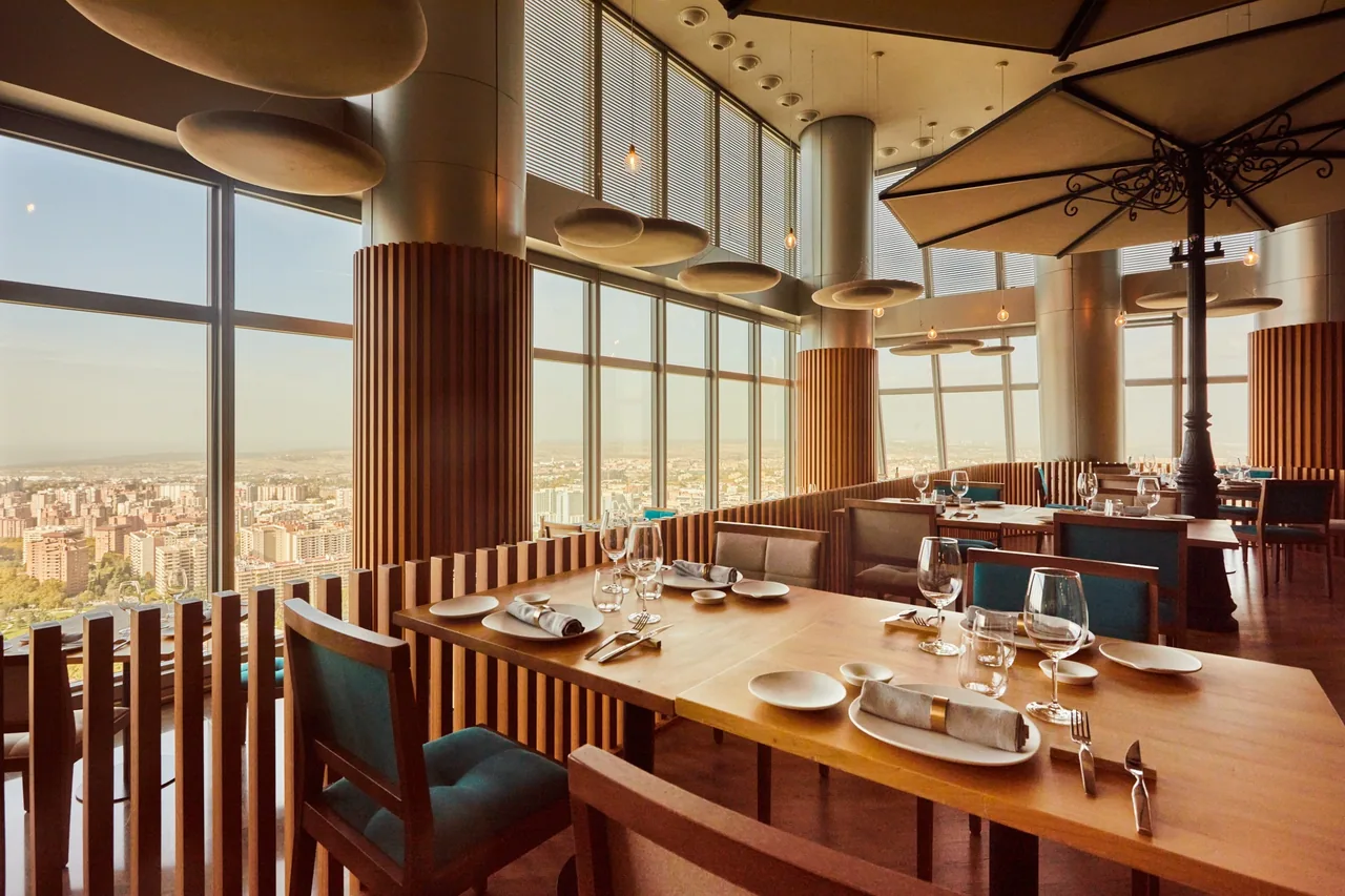 A dining room with wooden tables and teal chairs beside floor-to-ceiling windows looking out over the city