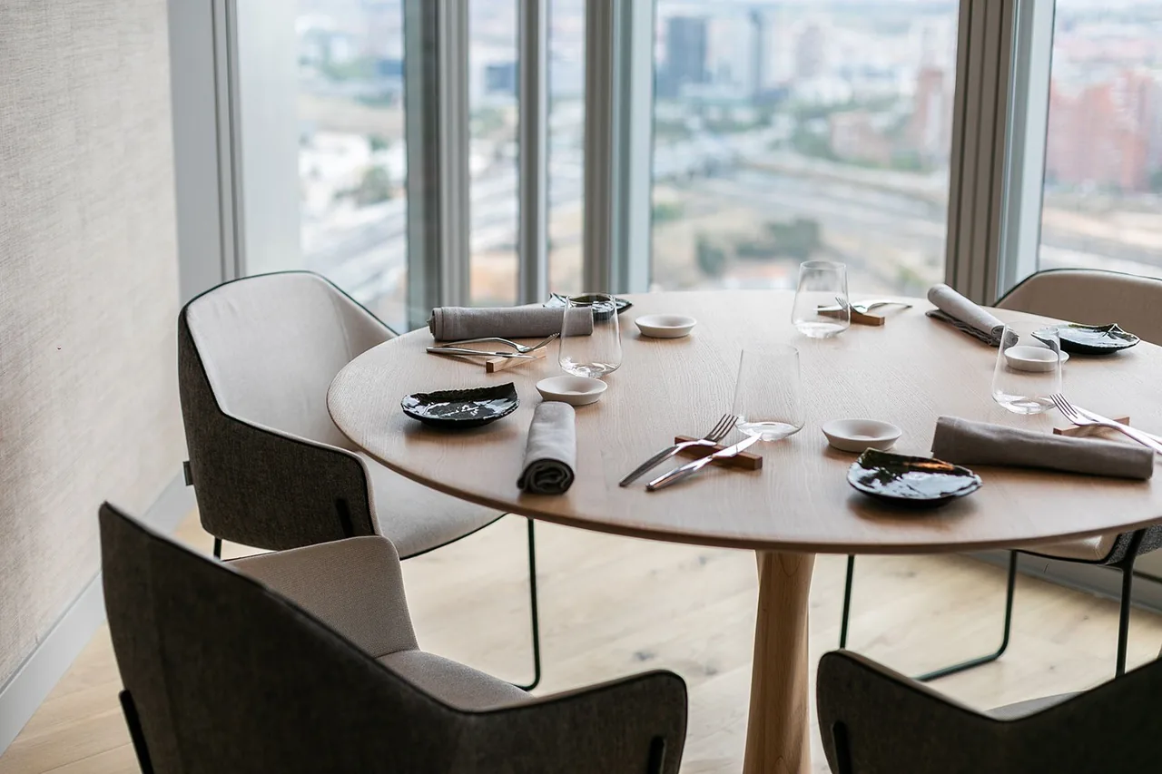 A round table set for dinner in front of a tall window with a city skyline beyond