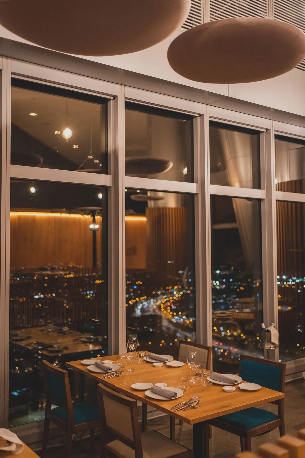 A wooden table by tall windows at night with city lights outside