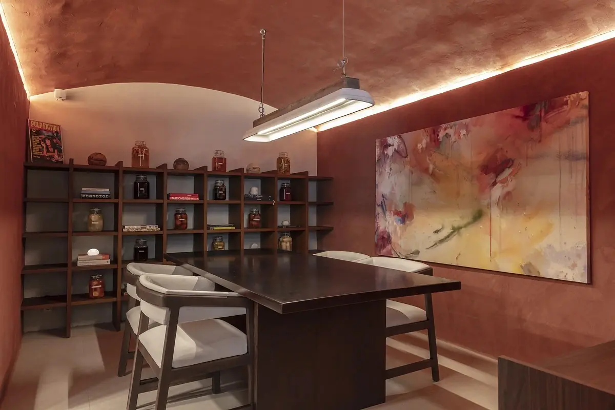 A small vaulted private dining room with terracotta plaster walls, a dark wood table and pale upholstered chairs, a grid of shelves holding preserving jars and books, and a large abstract painting in pinks and yellows