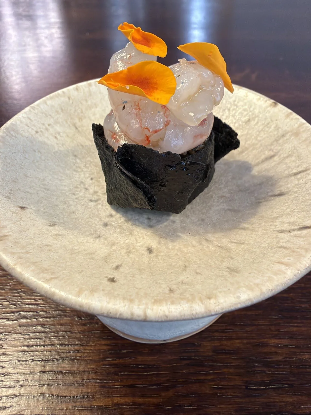A seafood bite in a black seaweed cup with an orange garnish, on a pale ceramic stand