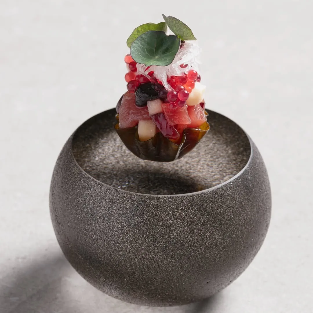 A colourful tartare of red and white pieces topped with green leaves and red pearls, held above a dark stone bowl