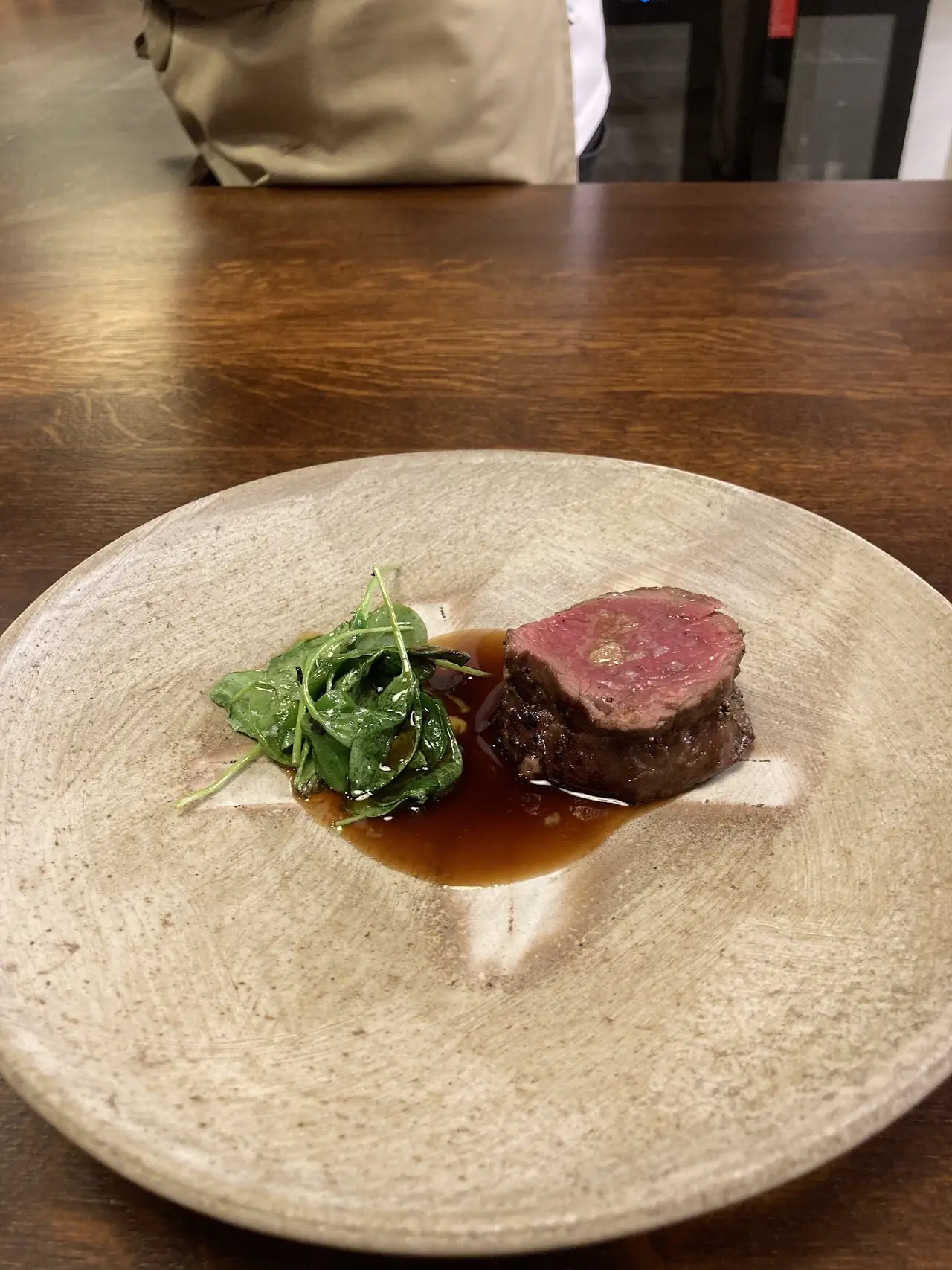 Sliced meat with a red interior, served with greens and a dark sauce on a textured plate.