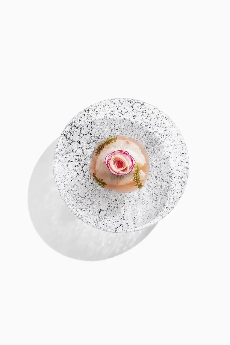 Small plated dish with a rolled fish garnish shaped like a rose, roe and herbs on a speckled ceramic plate