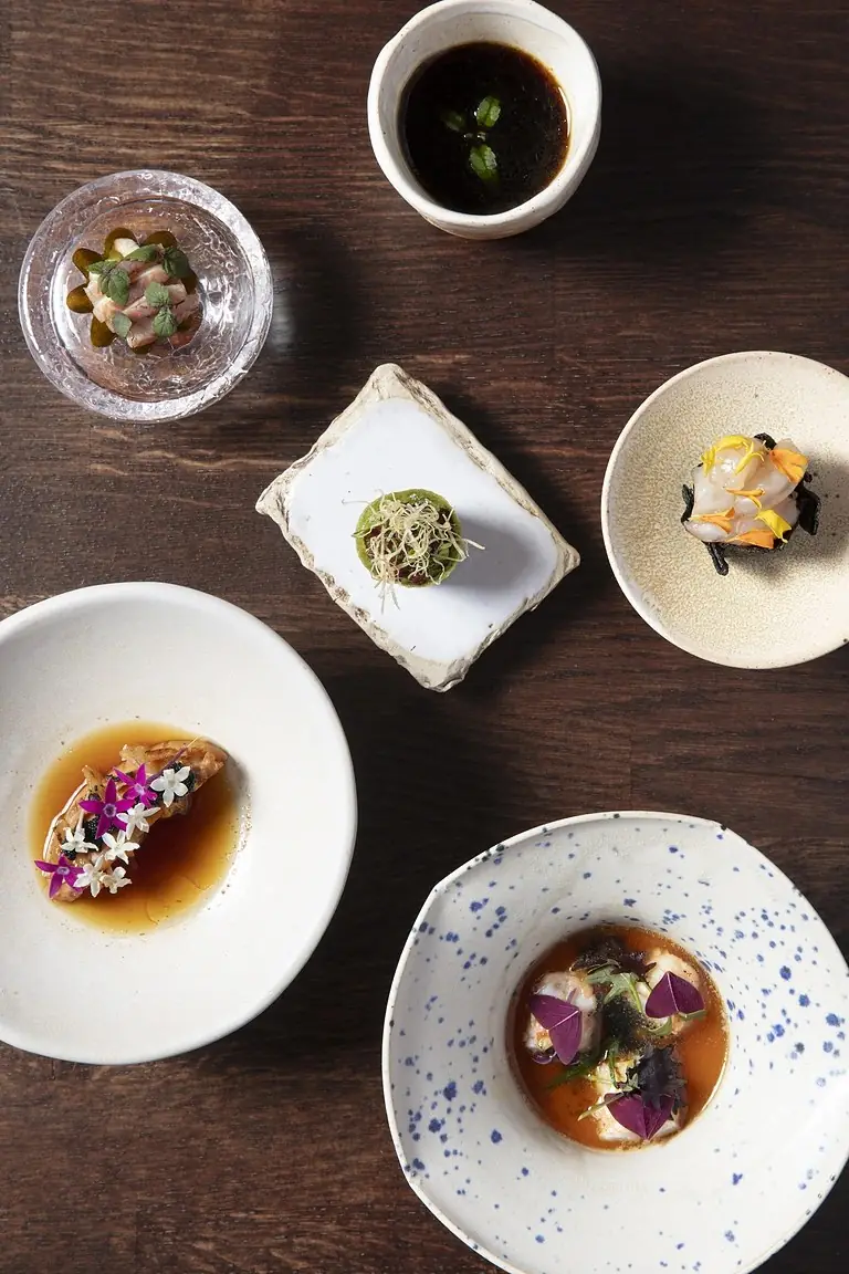 Overhead view of six tasting-menu courses on varied ceramic plates and bowls with broths, herbs and edible flowers