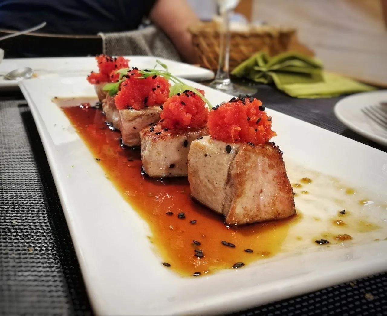 Slices of seared fish topped with red diced garnish on a long white plate with dark sauce